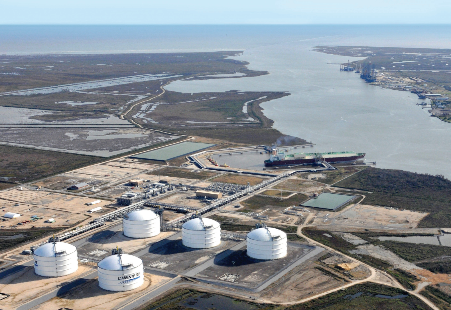 Aerial view of Cheniere’s Sabine Pass showing five large LNG tanks located near a river with an LNG tanker docked in an adjacent port.